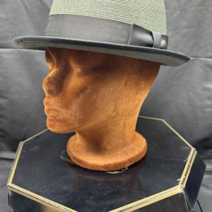 May include: A green fedora hat with a black band and brim, displayed on a brown mannequin head. The hat is sitting on a black box with the text "Cavanagh Hats". The hat has a classic, stylish design.