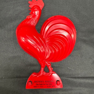 May include: Red plastic rooster figurine with a white base. The base has the text "BROWN'S MARKET Fine Foods for Fine Folks PHONE FE 2-8454 - ALCONY, OHIO".