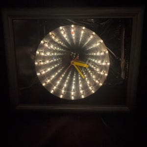 May include: A round clock with a spiral of small lights, set within a dark frame. The clock hands are yellow and red. The background is dark.