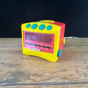 May include: A bright yellow and red RCA alarm clock radio with a digital display, four teal buttons on top, and two teal and yellow side knobs. The front panel features an AM/FM radio frequency scale with white text and a red background.