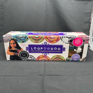 Könnte beinhalten: Eine farbenfrohe Schachtel mit einem Mädchen, das Armbänder aus bunten Schlaufen trägt. Auf der Schachtel steht "LOOPDEDOO" und "Gestalte deine eigenen Accessoires und fertige sie in wenigen Minuten an".