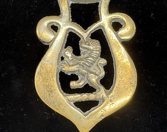 Puede incluir: Un antiguo adorno de latón para caballos con un diseño de león rampante. El latón tiene una forma decorativa con una figura central de león y detalles ornamentados. El fondo es de color oscuro.