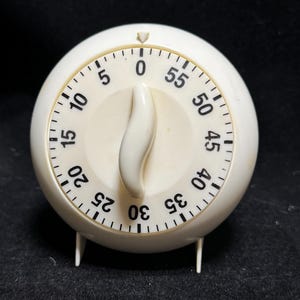 May include: A round, off-white kitchen timer with a central, curved rotating knob. The face features black printed numbers from 0 to 55 in increments of five, with tick marks for individual minutes. The device stands on two small legs.