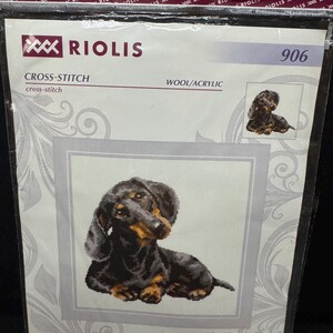 May include: A Riolis 906 cross-stitch kit featuring a black and tan dachshund. The packaging displays the text RIOLIS, 906, CROSS-STITCH, WOOL/ACRYLIC, and Dachshund. The finished design size is printed as 25 x 25 cm or 9 3/4" x 9 3/4".