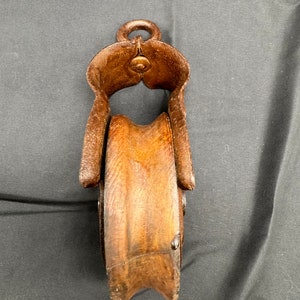 May include: A vintage wooden pulley with a metal frame. The pulley is brown and the frame is rusty. The pulley has a rope groove and a hook on top.