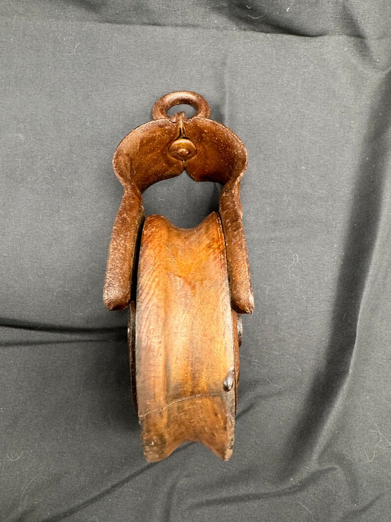 May include: A vintage wooden pulley with a metal frame. The pulley is brown and the frame is rusty. The pulley has a rope groove and a hook on top.