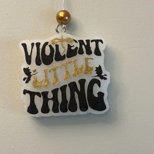 May include: A white, square-shaped air freshener with the words "VIOLENT LITTLE THING" in black and gold lettering. The air freshener is hanging from a string with a gold bead at the top. Decorative elements include small bat silhouettes.