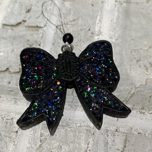 May include: A black glitter bow-shaped ornament with iridescent flecks. The bow has a decorative center and is suspended by a white string and a black bead. The ornament is likely for decoration.