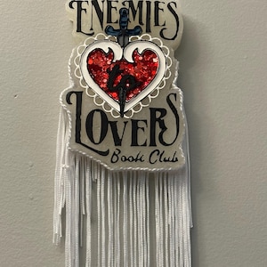 May include: A decorative hanging ornament with the words "ENEMIES" and "LOVERS Book Club" in black script. A red glitter heart pierced by a sword is centered between the words. White fringe hangs from the bottom.