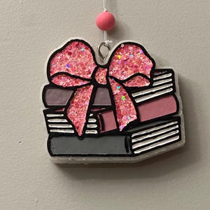May include: A decorative ornament featuring a stack of books topped with a pink glitter bow. The books are in shades of pink and gray, with black detailing. A pink bead hangs above the ornament.