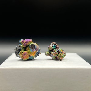 Könnte beinhalten: Zwei regenbogenfarbene, metallische, unregelmäßig geformte, steinartige Objekte mit einem Regenbogen aus Farben, darunter Grün, Blau, Lila und Rot, werden auf einer weißen Oberfläche präsentiert.