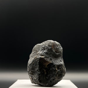 Può includere: Una roccia di colore grigio scuro, di forma irregolare, con una superficie ruvida e testurizzata. La roccia è appoggiata su una superficie bianca.