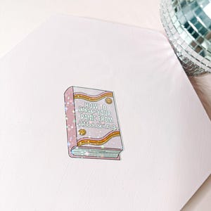 May include: A sticker in the shape of a book with the text "How to Stop Your Panic From Attacking". The book has a pink cover with yellow and orange stripes and small star accents. A disco ball is in the background.