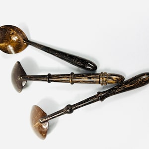 May include: Four handcrafted wooden kitchen utensils. The set includes a ladle and three different shaped spatulas. The utensils are made of dark brown wood with a natural grain pattern and a light brown scoop.