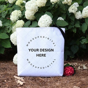 May include: White tote bag with black straps and the text "Mockups Digital" and "Your Design Here" in a circle. The bag is sitting on a bed of brown pine straw with white flowers in the background.