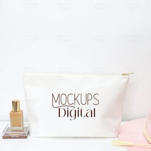 Op de afbeelding: Een witte ritszak met de tekst "MOCKUPS Digital" in bruin gedrukt op een witte achtergrond. De tas ligt op een wit oppervlak met een klein flesje vloeistof en een roze hoed.