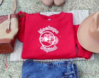 May include: A red crewneck sweatshirt with the words "Mockups Digital" and a play button graphic featuring cowboy hats. The sweatshirt is paired with blue jeans, a tan hat, and brown boots. A tan purse is also visible.