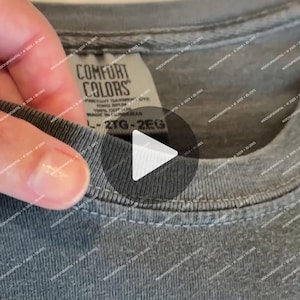 Könnte beinhalten: Nahaufnahme eines grauen Comfort Colors T-Shirt-Etiketts. Das Etikett zeigt "COMFORT COLORS" und enthält Informationen über die Färbung und das Material des Kleidungsstücks. Das Shirt ist ein gedämpftes Grau, und das Etikett wird von einer Hand gehalten.