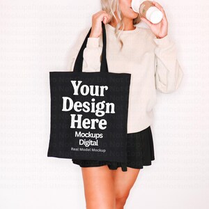 以下が含まれることがあります： 「Your Design Here」と「Mockups Digital Real Model Mockup」の白い文字が入った黒いトートバッグ。ベージュのスウェットシャツと黒いスカートを着た人がバッグを持っています。