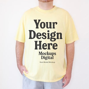 May include: A light yellow t-shirt with the text "Your Design Here" in a bold, black font. Below the text are the words "Mockups Digital" and "Real Model Mockup". The shirt is being worn by a person.