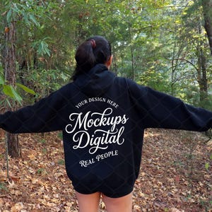May include: A black hooded sweatshirt featuring white text on the back that reads "YOUR DESIGN HERE Mockups Digital REAL PEOPLE" in a mix of serif and script fonts. The hoodie has long sleeves and a hood, shown from the back view.