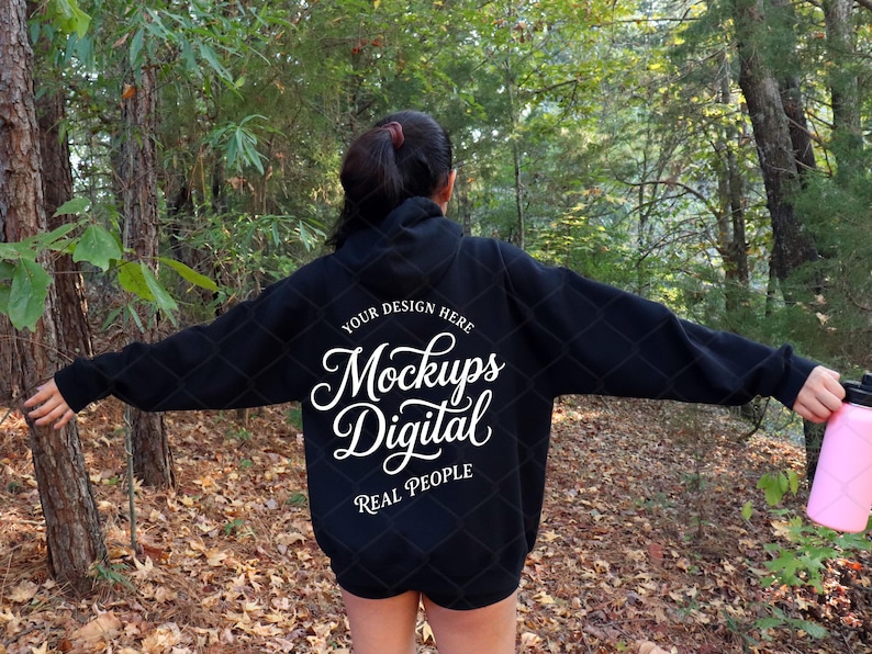 May include: A black hooded sweatshirt featuring white text on the back that reads "YOUR DESIGN HERE Mockups Digital REAL PEOPLE" in a mix of serif and script fonts. The hoodie has long sleeves and a hood, shown from the back view.