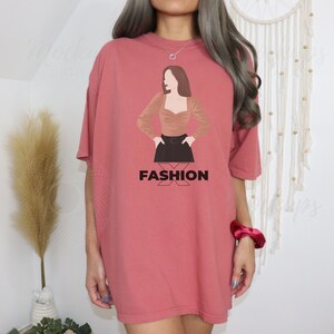 K&ouml;nnte beinhalten: Ein &uuml;berdimensioniertes T-Shirt in Korallenrosa mit einem Aufdruck einer Frau, die ein braunes Oberteil, einen schwarzen Rock und einen schwarzen G&uuml;rtel tr&auml;gt. Der Text "FASHION" ist unter dem Aufdruck gedruckt.