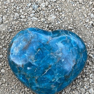 May include: A heart-shaped blue apatite crystal with a marbled pattern. The crystal is resting on a bed of small gray pebbles.