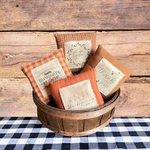 May include: Five orange and brown fabric throw pillows with Thanksgiving-themed text and designs. The pillows are arranged in a wooden basket on a black and white checkered tablecloth.