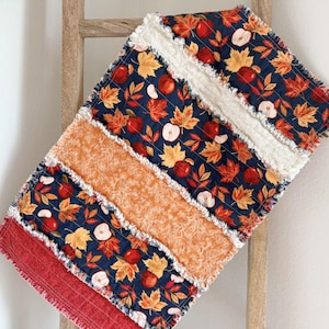 May include: A rag quilt featuring alternating strips of fabric. The pattern includes navy blue fabric with red apples and orange autumn leaves, a solid orange textured fabric, and a solid red fabric. The edges of the strips are frayed and white.