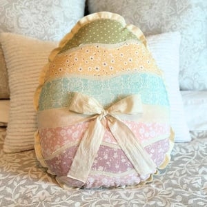 May include: A decorative Easter egg-shaped pillow with a patchwork design. The pillow features sections of pastel fabrics, including polka dots and floral patterns, and is tied with a cream-colored bow. The pillow is approximately 15 inches tall.