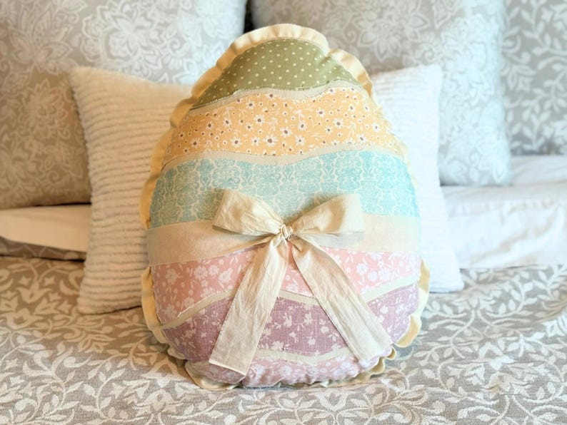 May include: A decorative Easter egg-shaped pillow with a patchwork design. The pillow features sections of pastel fabrics, including polka dots and floral patterns, and is tied with a cream-colored bow. The pillow is approximately 15 inches tall.