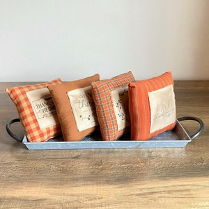 May include: Four decorative throw pillows with Thanksgiving-themed sayings on a light gray metal tray. The pillows are made of a plaid fabric in shades of orange and brown. The sayings on the pillows are "Baked Fresh, Thanksgivin' Pie Come On In", "Give Thanks with a Grateful Heart", "Happy Thanksgiving", and "Happy Thanksgiving".