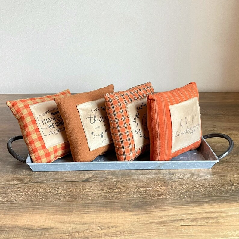 May include: Four decorative throw pillows with Thanksgiving-themed sayings on a light gray metal tray. The pillows are made of a plaid fabric in shades of orange and brown. The sayings on the pillows are "Baked Fresh, Thanksgivin' Pie Come On In", "Give Thanks with a Grateful Heart", "Happy Thanksgiving", and "Happy Thanksgiving".