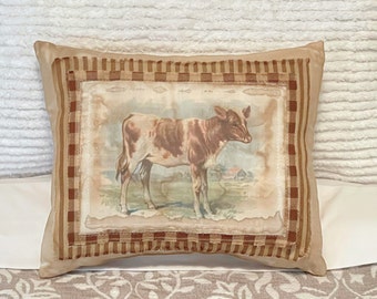 May include: A brown and white cow pillow with a gingham border. The pillow is on a white and brown patterned surface.