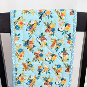 May include: A quilted table runner featuring a light blue background with a repeating pattern of colorful birds and floral arrangements. The birds are depicted in various colors, including red, blue, and yellow. The runner has a dotted border.