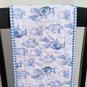 May include: A quilted table runner featuring a repeating pattern of blue chickens and egg baskets on a white background. The edges are trimmed with a blue and white gingham pattern. The runner is placed over the back of a black chair.