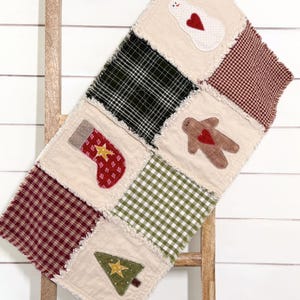May include: A rectangular rag quilt runner featuring alternating squares of cream fabric with appliqued holiday motifs including a snowman, a stocking, a gingerbread person, and a tree, alongside squares of red, green, and black plaid fabric.