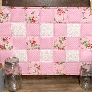 May include: A pink and white quilted placemat with a floral pattern. The placemat is made of squares of pink fabric with a diamond pattern, white fabric with a floral pattern, and pink fabric with red floral accents. The placemat is hanging from a wooden clothespin.