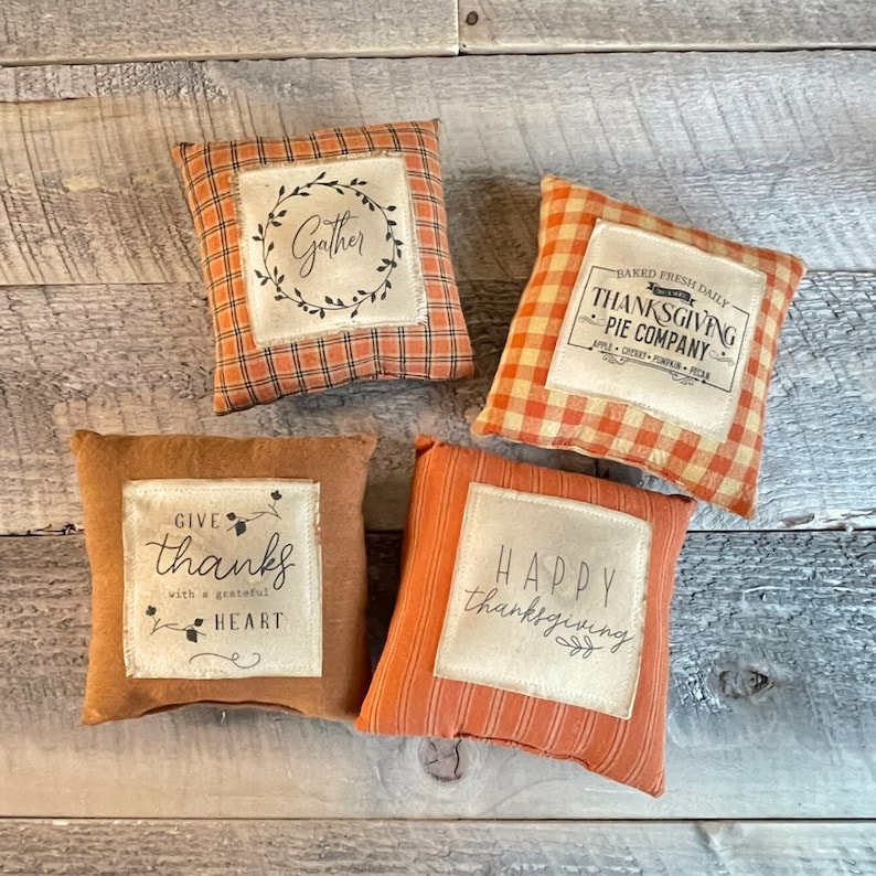 May include: Four decorative throw pillows with Thanksgiving-themed designs. The pillows are made of a brown and orange fabric with a plaid pattern. The pillows feature the following text: "Gather", "Baked Fresh Daily - Thanksgiving Pie Company", "Give Thanks with a Grateful Heart", and "Happy Thanksgiving".