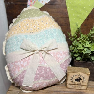 May include: An Easter egg-shaped pillow with a patchwork design in pastel colors, including pink, yellow, and blue, is tied with a cream-colored bow. A small wooden block with a rabbit silhouette and the text "Happy Easter" is also visible.