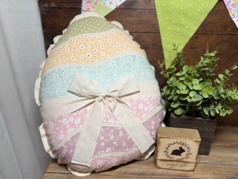 May include: An Easter egg-shaped pillow with a patchwork design in pastel colors, including pink, yellow, and blue, is tied with a cream-colored bow. A small wooden block with a rabbit silhouette and the text "Happy Easter" is also visible.