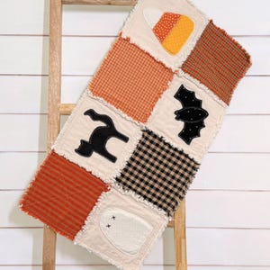 May include: A rectangular rag quilt featuring a patchwork design with squares of orange plaid, black and white gingham, and cream fabric. Appliqué motifs include a candy corn, a black cat, a black bat, and a white ghost with stitched eyes.