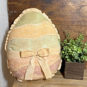 May include: An Easter egg-shaped pillow with a ruffled edge. The pillow features horizontal stripes in pastel colors, including green, yellow, and pink, with a bow at the center. A small plant in a wooden cube sits nearby.