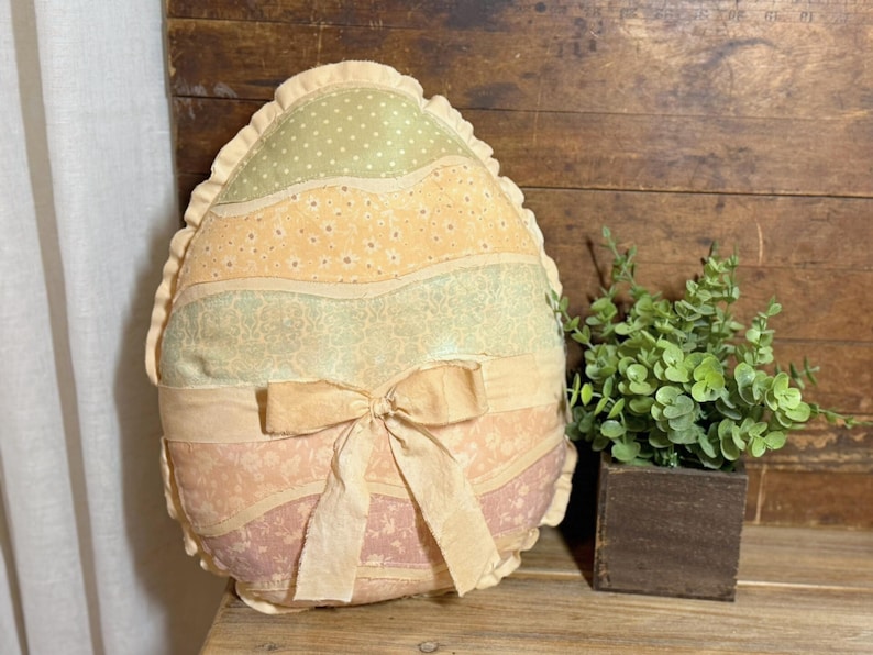 May include: An Easter egg-shaped pillow with a ruffled edge. The pillow features horizontal stripes in pastel colors, including green, yellow, and pink, with a bow at the center. A small plant in a wooden cube sits nearby.