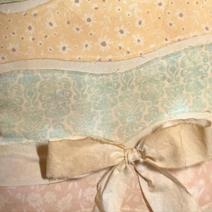May include: Close-up of a decorative pillow with a layered design. The top layer is yellow with small white flowers, followed by a light blue patterned layer, and a pink floral layer. A cream-colored bow is tied at the bottom.