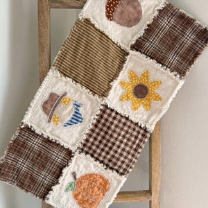 May include: A patchwork rag quilt featuring alternating squares of brown plaid, striped, and solid fabrics. Appliqué designs include a brown acorn, a yellow sunflower, a brown hat with blue accents, and an orange pumpkin on cream-colored squares.