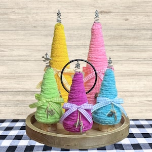 May include: A collection of five colorful, handmade decorative trees. The trees are cone-shaped, wrapped in textured yarn in yellow, pink, green, purple, and blue. Each tree is adorned with a ribbon bow and a metal charm, and topped with a silver spiral. They sit on a wooden tray.