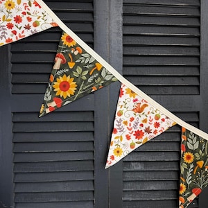 May include: A festive banner with triangular flags. The flags feature autumn-themed patterns, including sunflowers, mushrooms, and squirrels, set against white and dark green backgrounds. The banner is strung on a cream-colored ribbon, perfect for fall decor.