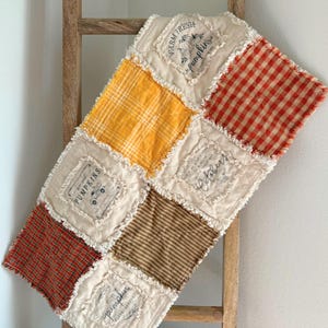 May include: A rag quilt featuring alternating squares of cream fabric and patterned fabric in orange, yellow, and brown. The cream squares display printed text and graphics including Farm Fresh Pumpkins, October, and Pumpkins. The edges are frayed.